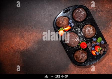 Schokoladen-Muffins mit Beeren. Muffin-Pfanne mit hausgemachten süßen, dunklen Schokoladen-Cupcakes mit Kakaosoße, Zuckerpulver und frischen Erdbeeren, Blaubeeren Stockfoto