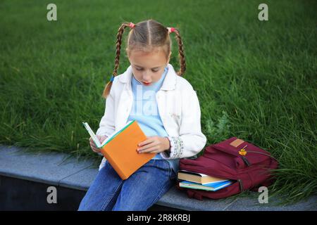 Süßes kleines Mädchen mit Rucksack, das Lehrbuch auf der City Street liest Stockfoto