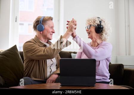 Ältere Männer und Frauen geben sich gegenseitig High Five, während sie im Wohnzimmer vor dem digitalen Tablet sitzen Stockfoto