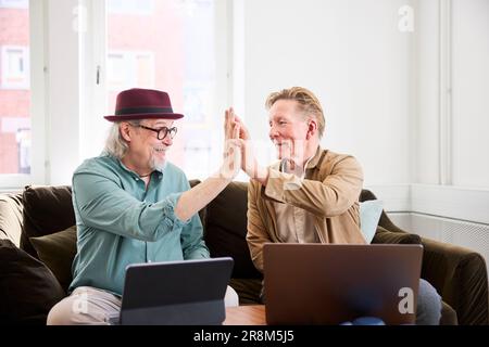 Ältere Männer geben sich gegenseitig High Five, während sie im Wohnzimmer vor dem digitalen Tablet sitzen Stockfoto