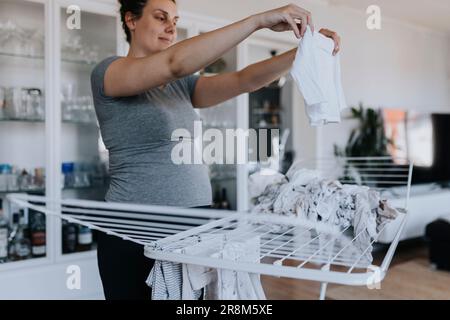 Schwangere Frau, die Hausarbeit macht und Babykleidung an einen Trockenständer hängt Stockfoto