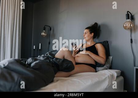 Schwangere Frau, die sich im Bett entspannt und telefoniert Stockfoto
