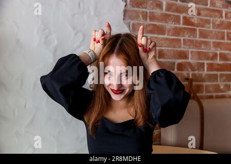 Das Mädchen im Café zeigt ein komisches Gesicht. Ein Mädchen mit roten Haaren und einem schwarzen Kleid in einem Café zeigt ein lustiges Gesicht Stockfoto