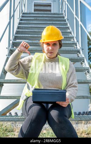 Ein Bauarbeiter mit Schutzhelm sitzt auf einer Leiter, während er einen Laptop für die Arbeit benutzt Stockfoto