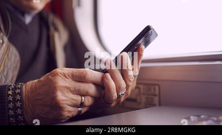 Nahaufnahme der Hände eines Seniors, der in einem Zugwagen sitzt und ein Smartphone benutzt Stockfoto