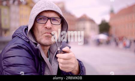 Porträt eines erwachsenen Mannes mit Brille, der in der Kapuze auf dem Platz sitzt und eine Tabakpfeife raucht, auf dem Palastplatz, Warschauer Altstadt Stockfoto