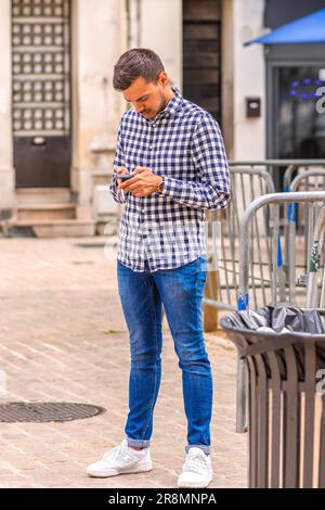 Junger Mann in kariertem Hemd und Jeans, der auf der Straße der Stadt steht und sein Handy überprüft - Tours, Indre-et-Loire (37), Frankreich. Stockfoto