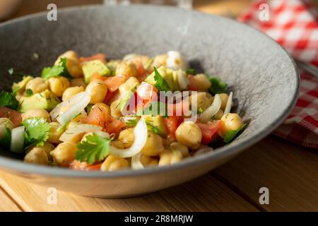 45°-Blick auf einen Kichererbsensalat mit Avocado, Tomaten, Koriander und Zwiebeln, serviert auf einem grauen Gericht mit einem roten karierten Tuch auf einem Holztisch. Stockfoto