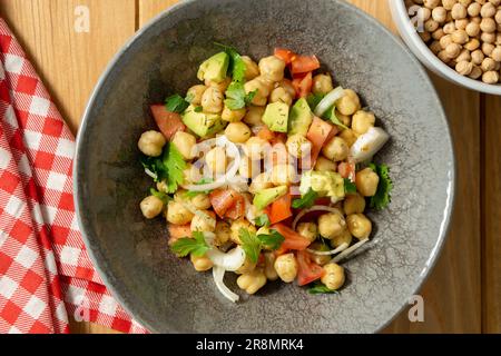 Von oben aus haben Sie einen Blick auf einen Kichererbsensalat mit Avocado, Tomaten, Koriander und Zwiebeln, serviert auf einem grauen Gericht mit einem roten Tuch, Kichererbsen und einem Holztisch. Stockfoto