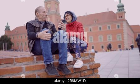 Ältere Touristen, die auf ihr Handy schauen, sitzen im historischen Zentrum einer alten europäischen Stadt und unterhalten sich miteinander. Palast Stockfoto