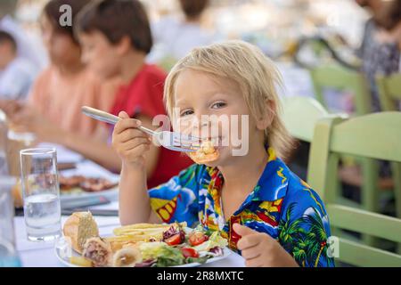 Süßes Kind, das im Sommer im Restaurant im Freien sitzt und Meeresfrüchte, Garnelen, Calamari, Tintenfisch und pommes frites isst Stockfoto