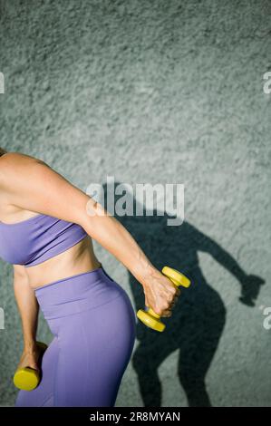 Seitenansicht einer sportlichen Frau in leuchtender Aktivbekleidung, die Trizeps, Kickbacks und Kurzhanteln in hellem Licht an der grauen Wand macht Stockfoto