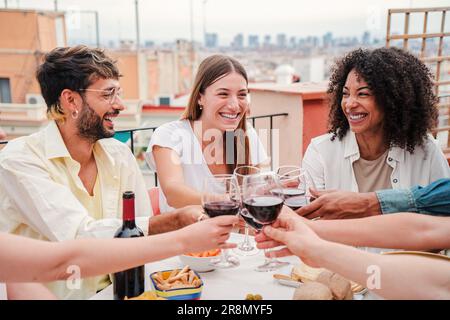 Eine Gruppe glücklicher Freunde, die ein Glas Rotwein trinken und den Moment genießen, ein Aperitif auf dem Dach des Hauses. Lächelnde junge Leute, die anstoßen, trinken und reden, während sie Spaß miteinander haben. Hochwertiges Foto Stockfoto