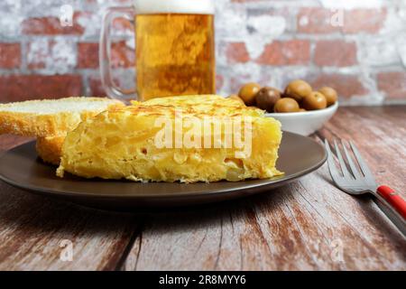 Typisch spanisches Kartoffelomelett, frisch zubereitet auf einem Holztisch mit einem frischen Krug aus Bier, Besteck, Brot und Oliven Stockfoto