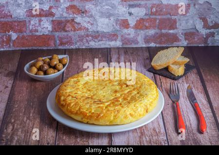 Typisch spanisches Kartoffelomelett, frisch zubereitet auf einem Holztisch mit Besteck, Brot und Oliven Stockfoto