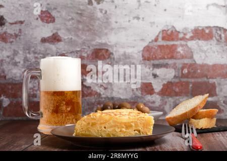 Typisch spanisches Kartoffelomelett, frisch zubereitet auf einem Holztisch mit einem frischen Krug aus Bier, Besteck, Brot und Oliven Stockfoto