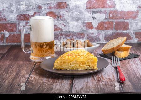 Typisch spanisches Kartoffelomelett, frisch zubereitet auf einem Holztisch mit einem frischen Krug aus Bier, Besteck, Brot und Oliven Stockfoto