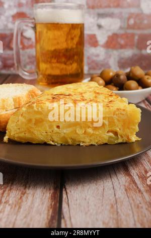 Typisch spanisches Kartoffelomelett, frisch zubereitet auf einem Holztisch mit einem frischen Krug aus Bier, Besteck, Brot und Oliven Stockfoto