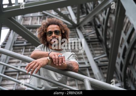 Porträt eines jungen Mannes mit Afro-Haaren, der eine Sonnenbrille auf der Treppe in der Stadt trägt, urbane Mode cooles Konzept eines Hipster-Mannes mit Stil Stockfoto