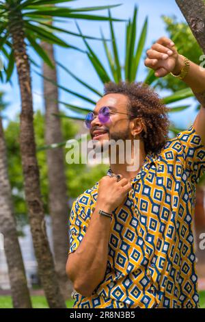 Porträt eines afrohaarigen Mannes im Sommerurlaub neben ein paar Palmen am Strand, der lächelt und nach links auf eine Palme lehnt. Reise- und Stockfoto