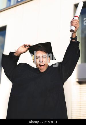 Ein fröhlicher, lächelnder Absolvent, der ein Kleid trägt und sein Diplom hochzieht Stockfoto