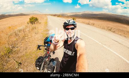 Ein weißer Mann steht am Fahrrad allein und fährt durch die ruhige Landschaft, voll mit Taschen im Freien, im Sommer zeigt die Hitze Daumen hoch. Kreative ec Stockfoto