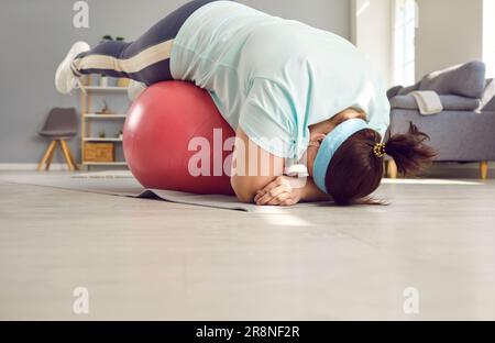 Lustiges, fettes, übergewichtiges Mädchen, das Sportkleidung trägt und zu Hause Sportübungen mit fit Ball macht. Stockfoto