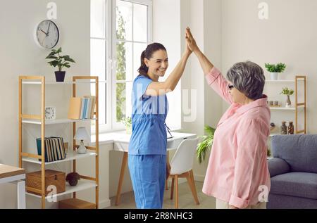 Lustige und zufriedene Seniorin gibt High Five an junge Krankenschwester, die sie zu Hause besucht hat. Stockfoto