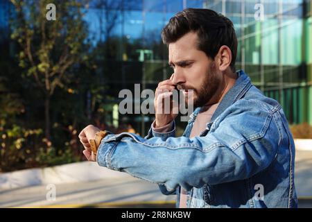 Emotionaler Mann, der auf einem Smartphone spricht und auf eine Uhr in der Nähe eines Gebäudes schaut. Das Konzept der Verspätung Stockfoto