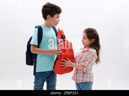 Ein junger Junge mit einem orangefarbenen Rucksack seiner jüngeren Schwester, bereit für die Schule, isoliert vor weißem Hintergrund. Stockfoto