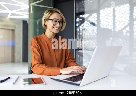 Glückliche und lächelnde Geschäftsfrau, die auf dem Laptop tippt, Büroangestellter mit Brille, zufrieden mit den erzielten Ergebnissen, bei der Arbeit im Bürogebäude Stockfoto