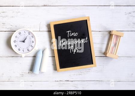 Blick von oben auf Kreide, Tischuhr, Sanduhr und Tafel mit „ZEITMANAGEMENT-TIPPS“ auf weißem Holzhintergrund. Stockfoto