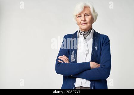 Eine seriöse ältere Frau in modischer Kleidung, die die Arme auf der Brust kreuzt und im Studio in die Kamera schaut Stockfoto