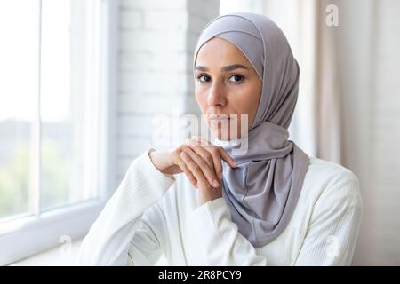 Nahaufnahme. Porträt einer jungen, wunderschönen muslimischen Frau in einem weißen Hidschab, der drinnen am Fenster steht. Er schaut selbstbewusst und ernsthaft in die Kamera. Stockfoto