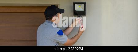 Bild eines Elektrikers auf einer Scherenleiter, der mit seinen Werkzeugen einen Scheinwerfer an einem Haus installiert. Beleuchtung der dunkelsten Teile des Hauses. Banner Stockfoto