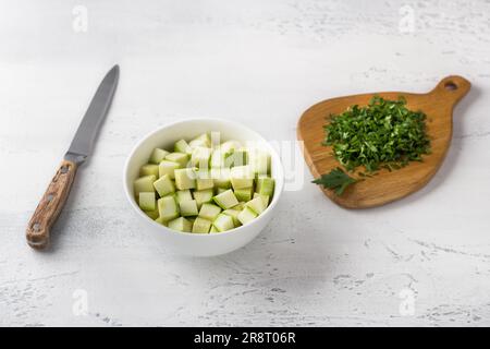 Eine Schüssel gewürfelter Zucchini, ein Holzbrett mit Kräutern und einem Messer auf hellblauem Hintergrund. Köstliches hausgemachtes Frühstück. Stockfoto