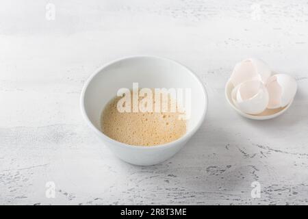 Eine weiße Schüssel mit verprügelten Eiern und ein Teller mit einer Schale auf hellblauem Hintergrund. Ein köstliches hausgemachtes Omelette. Stockfoto