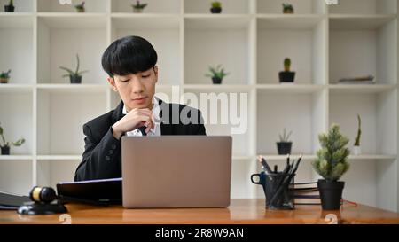 Ein kluger und professioneller männlicher asiatischer Anwalt oder Rechtsberater in einem formellen Rechtsstreit konzentriert sich auf seine Arbeit auf seinem Laptop und liest ein Gerichtsdokument auf seinem La Stockfoto