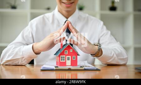 Nahaufnahme eines professionellen männlichen Immobilienmaklers oder Versicherungsmaklers, der eine Geste zum Schutz der Hände über ein Hausmodell auf einem Tisch macht. pro Stockfoto