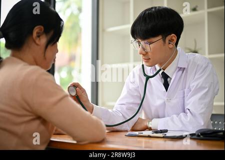 Ein fokussierter und professioneller Millenialasiatischer männlicher Arzt verwendet ein Stethoskop, um den Herzschlag einer Patientin im Krankenhaus zu untersuchen Stockfoto