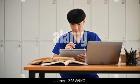 Ein intelligenter und fokussierter junger asiatischer Medizinstudent in Peelings liest eine Online-medizinische Zeitung auf seinem Tablet an einem Tisch. Stockfoto