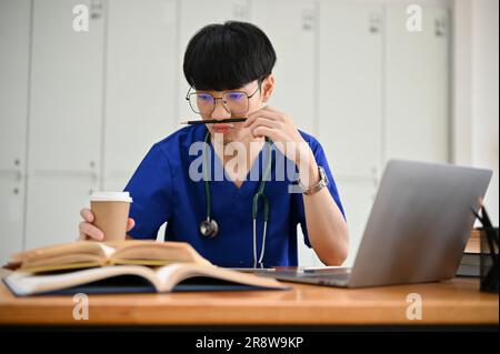 Ein glücklicher und gutaussehender junger asiatischer Medizinstudent in Peelings konzentriert sich auf seine medizinische Forschung, liest ein Buch und trinkt Kaffee an einem Tisch in seinem Stockfoto
