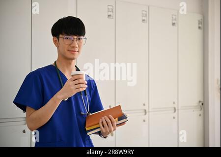 Ein Porträt eines gutaussehenden und positiven jungen asiatischen Medizinstudenten oder Arztes in Peelings steht im Krankenhausflur mit einer Kaffeetasse und Büchern Stockfoto