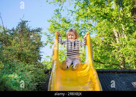 Fröhlicher, süßer Junge, der Spaß auf einer gelben Rutsche im Park hat, sonniger Sommertag auf dem Kinderspielplatz. Selektiver Fokus Stockfoto