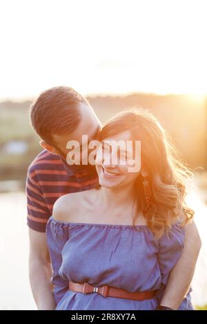 Mann und schwangere Frau in Kleidern umarmen sich im Sommer bei Sonnenuntergang Stockfoto