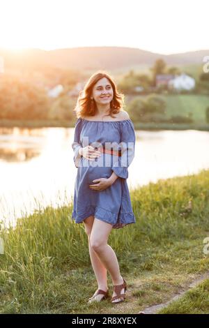 Schwangere Frau in blauem Kleid hält ihren Bauch unter Sonnenstrahlen Stockfoto