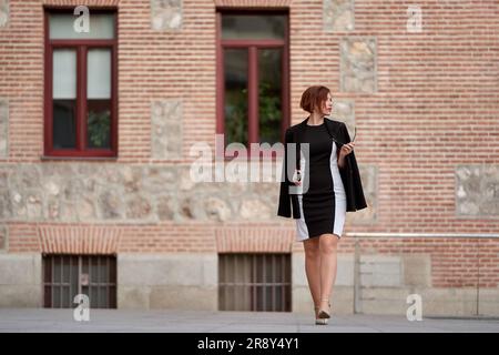 Rothaarige Geschäftsfrau, die in einem eleganten Kleid und einer Sonnenbrille spaziert Stockfoto