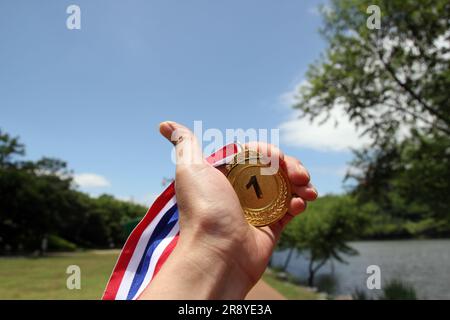 Verwischte Frauenhände, die Goldmedaillen mit thailändischem Band vor blauem Himmel halten, um Erfolg im Sport oder Geschäft zu zeigen, Gewinner erfolgreich Stockfoto