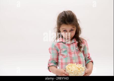 Ein hübsches Mädchen im stilvollen pinkfarbenen und blauen Karohemd, das eine Schüssel mit köstlichem Popcorn hält, isoliert auf weißem Hintergrund Stockfoto