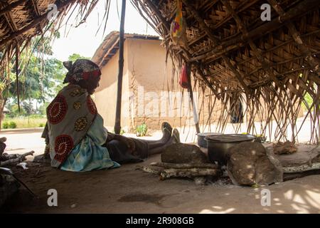 Eine alte Dame aus dem Makonde-Stamm sitzt in der primitiven Küche und bereitet lokale traditionelle Mahlzeit zu. Einfacher Metalltopf auf Feuer zwischen Steinen Stockfoto